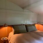 Lit double avec linge dans des tons terre cuite et oreillers assortis, encadré par deux lampes suspendues diffusant une lumière chaude, dans une chambre mansardée aux murs blancs et ambiance apaisante