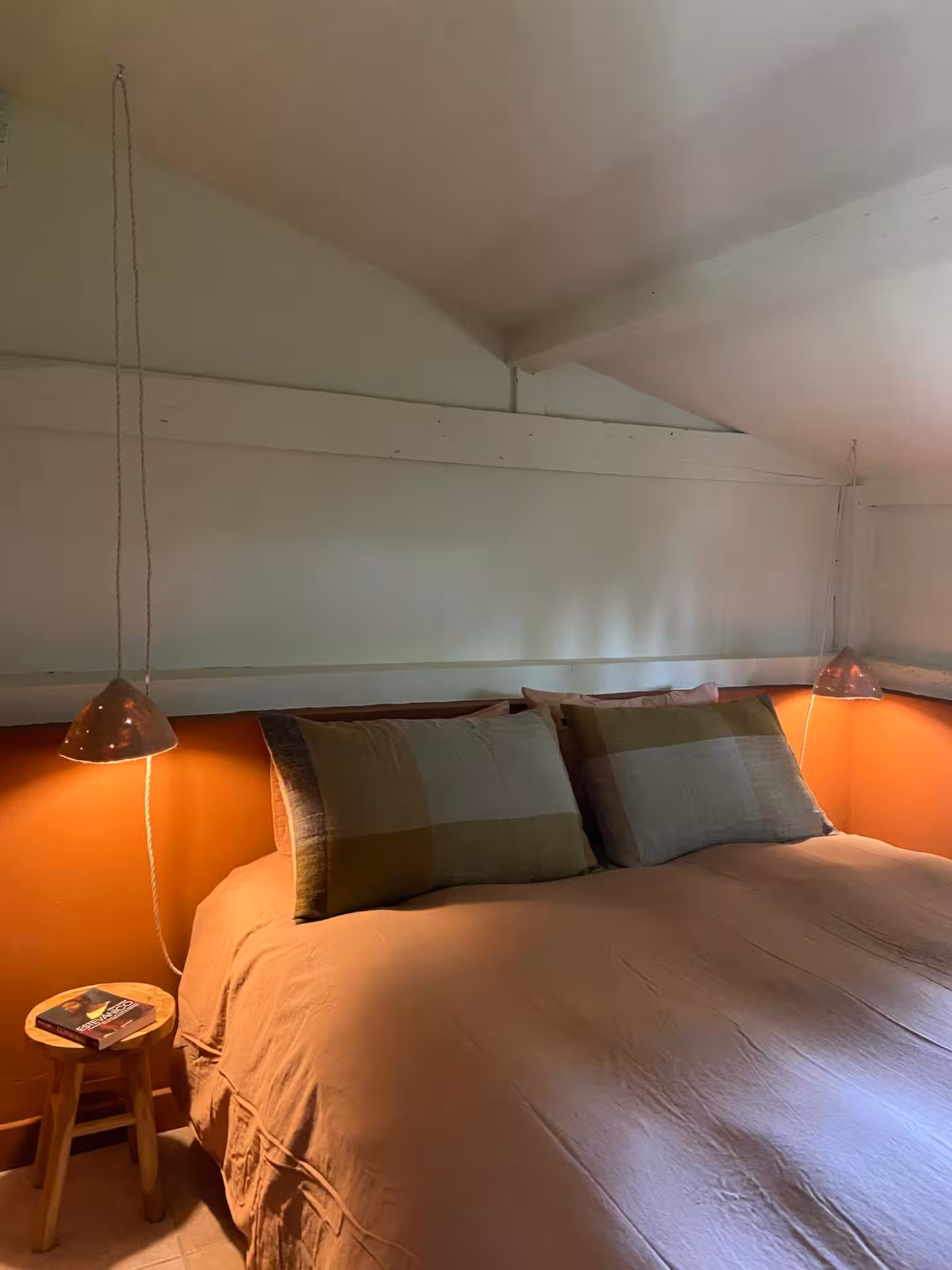 Lit double avec linge dans des tons terre cuite et oreillers assortis, encadré par deux lampes suspendues diffusant une lumière chaude, dans une chambre mansardée aux murs blancs et ambiance apaisante