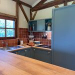 Cuisine chaleureuse au style rustique avec meubles bas bleu mat, crédence en carrelage brun, plan de travail en bois clair et poutres apparentes sous plafond