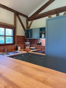 Cuisine chaleureuse au style rustique avec meubles bas bleu mat, crédence en carrelage brun, plan de travail en bois clair et poutres apparentes sous plafond