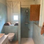 Salle de bain lumineuse au style rustique avec douche à l’italienne, toilettes suspendues, murs en petits carreaux bleu clair et poutres apparentes peintes en blanc