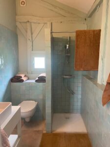 Salle de bain lumineuse au style rustique avec douche à l’italienne, toilettes suspendues, murs en petits carreaux bleu clair et poutres apparentes peintes en blanc