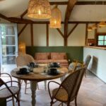 Salle à manger et salon ouverts avec poutres en bois apparentes, mobilier en rotin et bois clair, suspensions en osier, canapé en lin, et cuisine ouverte à l’arrière-plan, le tout baigné de lumière naturelle