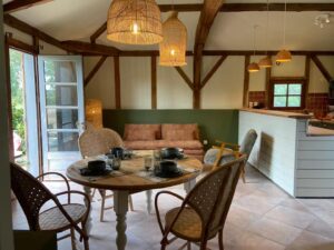 Salle à manger et salon ouverts avec poutres en bois apparentes, mobilier en rotin et bois clair, suspensions en osier, canapé en lin, et cuisine ouverte à l’arrière-plan, le tout baigné de lumière naturelle