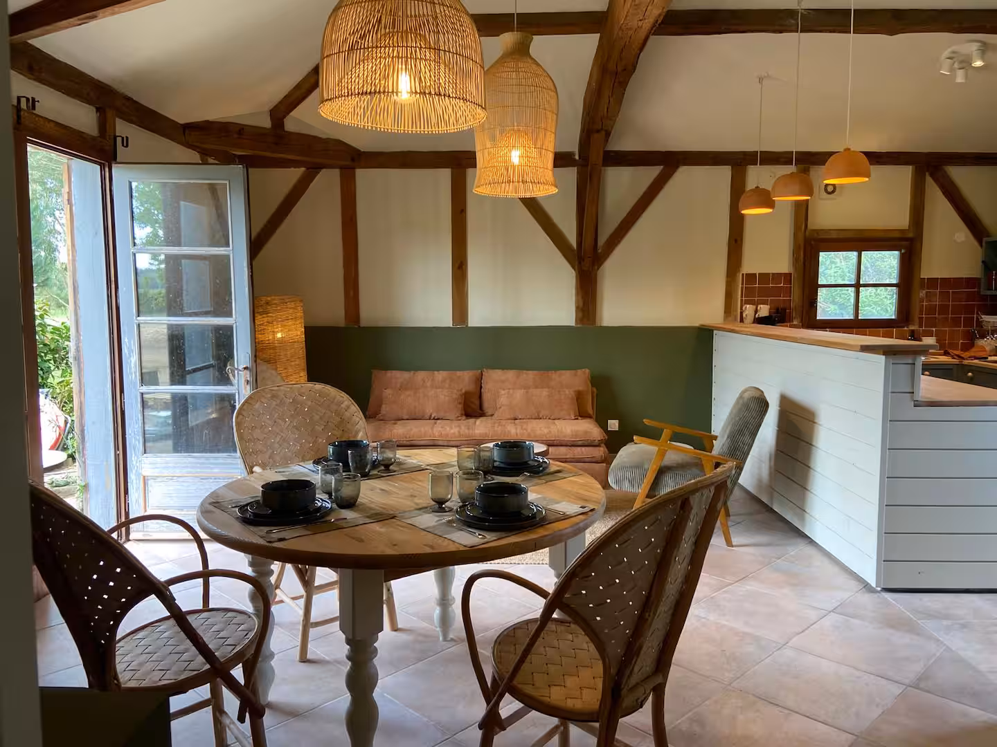 Salle à manger et salon ouverts avec poutres en bois apparentes, mobilier en rotin et bois clair, suspensions en osier, canapé en lin, et cuisine ouverte à l’arrière-plan, le tout baigné de lumière naturelle