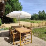 Terrasse en béton accolée à une maison, aménagée avec une table et des chaises en bois sous un parasol blanc à franges, donnant sur un espace naturel bordé de clôtures en bois et de végétation