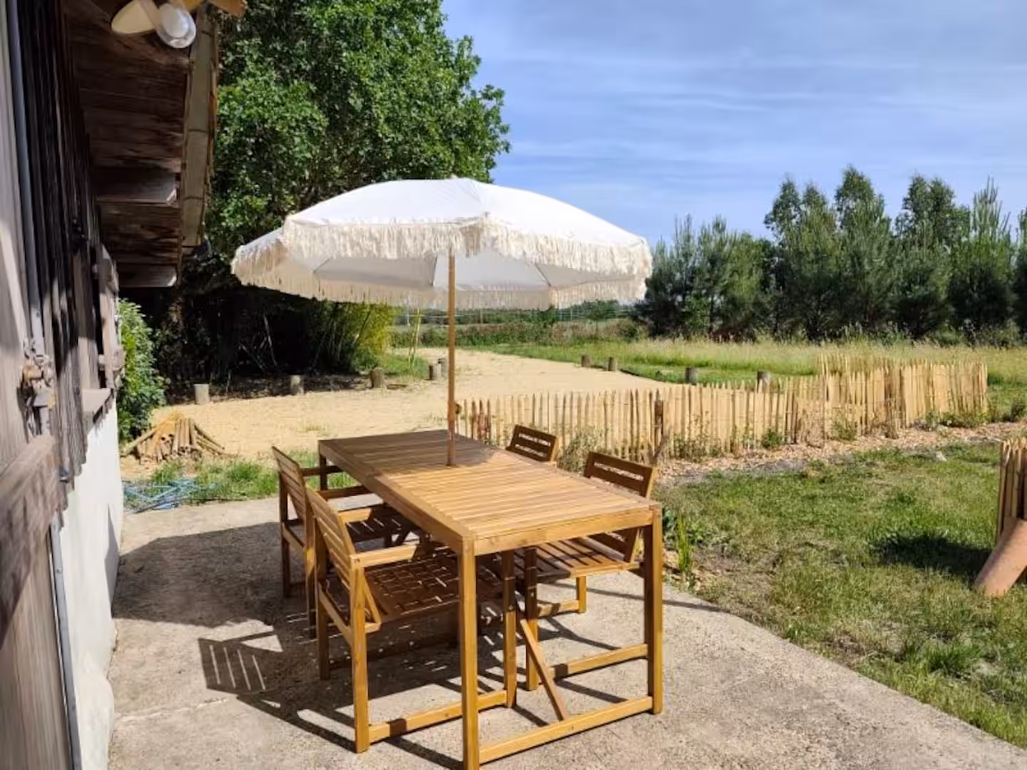 Terrasse en béton accolée à une maison, aménagée avec une table et des chaises en bois sous un parasol blanc à franges, donnant sur un espace naturel bordé de clôtures en bois et de végétation